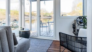 Une pièce comprenant un canapé, une petite table avec des plantes, une chaise noire et une vue sur une étendue d’eau, visible à travers de grandes portes coulissantes.