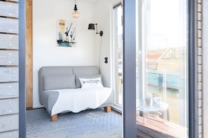 Une pièce moderne avec un canapé gris, une petite table ronde, une lampe fixée au mur et une grande fenêtre.