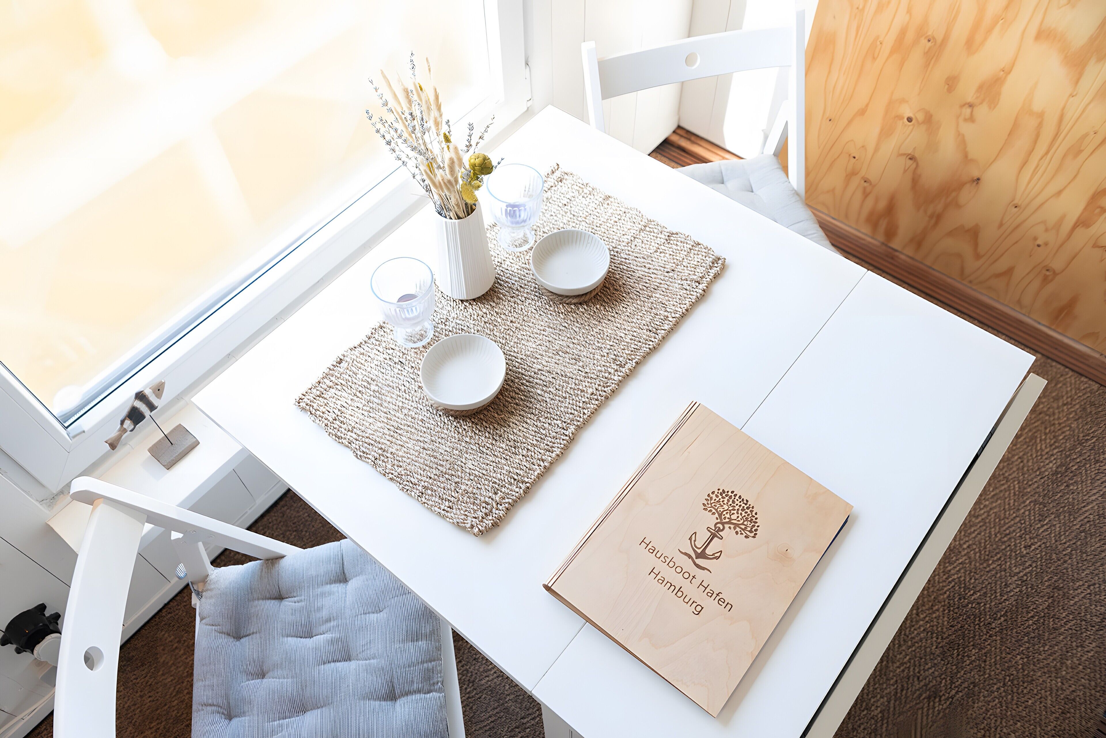 Una mesa blanca con un camino de mesa, dos cuencos, un florero con flores secas y una señal de madera con texto.