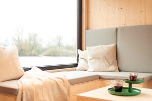 Un coin salon confortable avec un banc rembourré, une petite table avec une bougie et une fenêtre donnant sur les arbres.