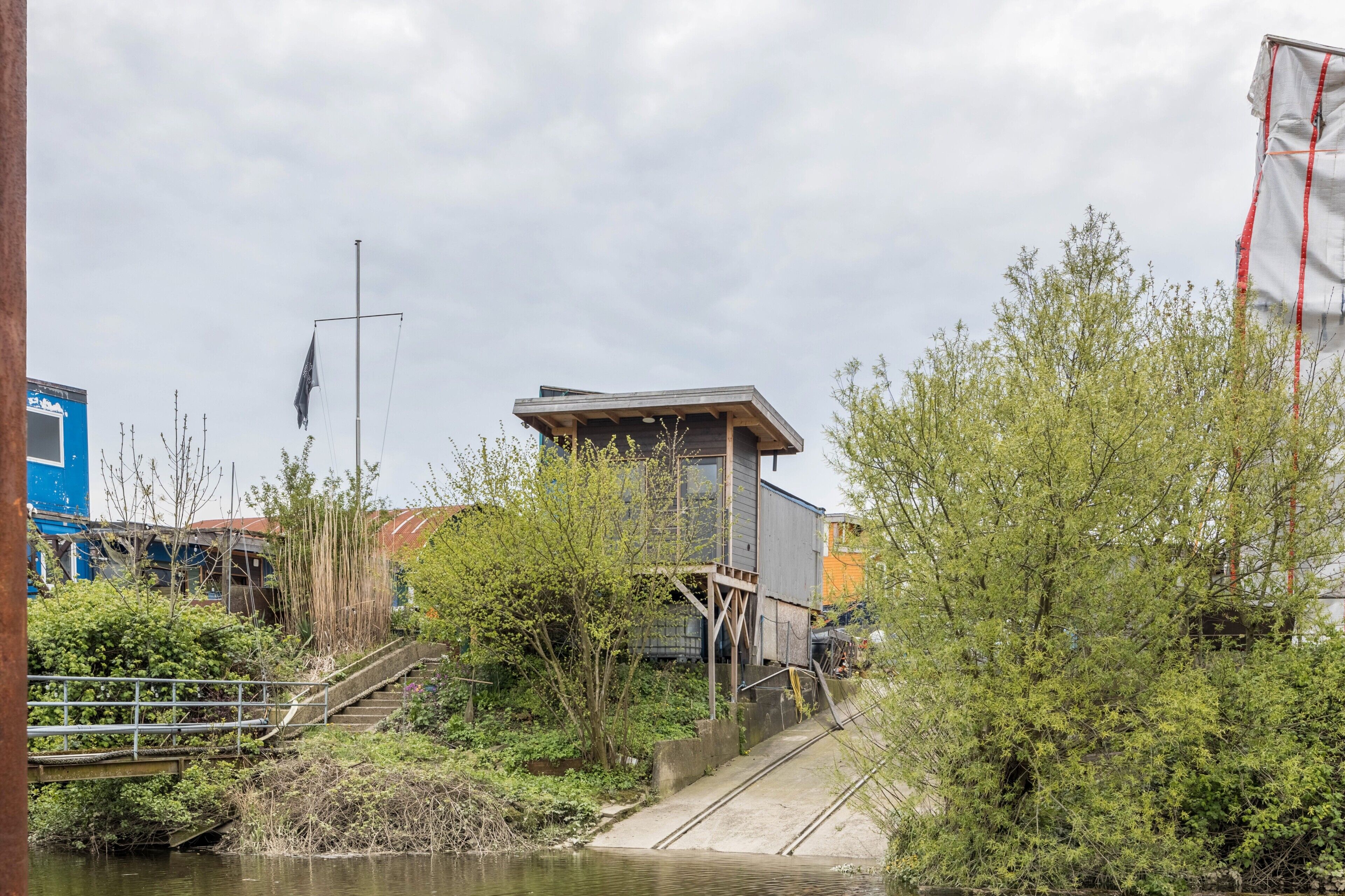 A riverside building with a staircase leading to a platform, surrounded by greenery and a bridge.