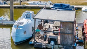 A floating houseboat docked at a marina with a small deck area, a water pump, and a lifebuoy.
