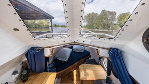 A boat interior with a wooden table, a bench, and two windows offering views of a dock and trees.
