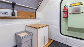 Een compacte kitchenette met een prullenbak, een klein kastje en een brandblusser aan de muur.