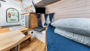 A compact RV interior with a bed, a small kitchenette, a mounted TV, and a door with a fire extinguisher.