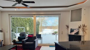 Una sala de estar con televisor, ventilador de techo, chimenea y un amplio ventanal corredera que da a un área exterior nevada.