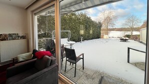 Una habitación con un amplio ventanal corredera que da a un área exterior nevada con un patio, una silla y una mesa.