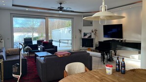 Una sala de estar moderna con un ventanal grande, un ventilador de techo, una chimenea, un piano y un televisor.