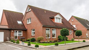 Ein Wohnhaus aus rotem Backstein mit einem geneigten Dach, einer Veranda und einem gepflegten Garten mit verschiedenen Sträuchern und einer Rasenfläche.