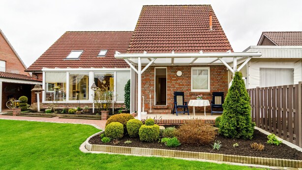 Ein Haus mit überdachter Terrasse, Ziegelaußenwand und gepflegtem Garten mit verschiedenen Pflanzen und Rasenfläche.