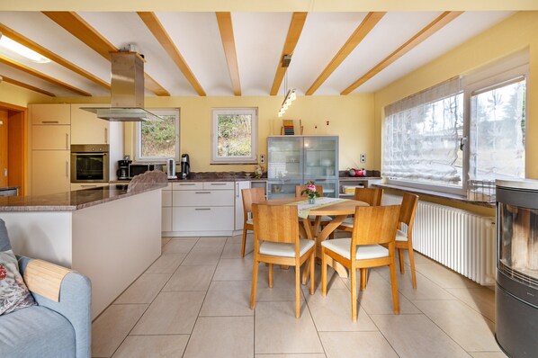 A modern kitchen with a central island, wooden ceiling beams, and a dining area with chairs and a table.