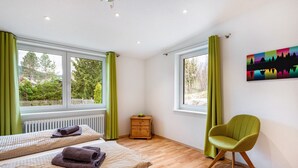 Una habitación con dos camas, piso de madera, una silla verde y una ventana con vista a árboles.
