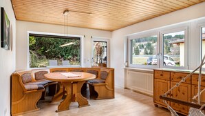 Un comedor con mesa y bancos de madera, ventanas amplias con vistas a la vegetación y techo de madera.