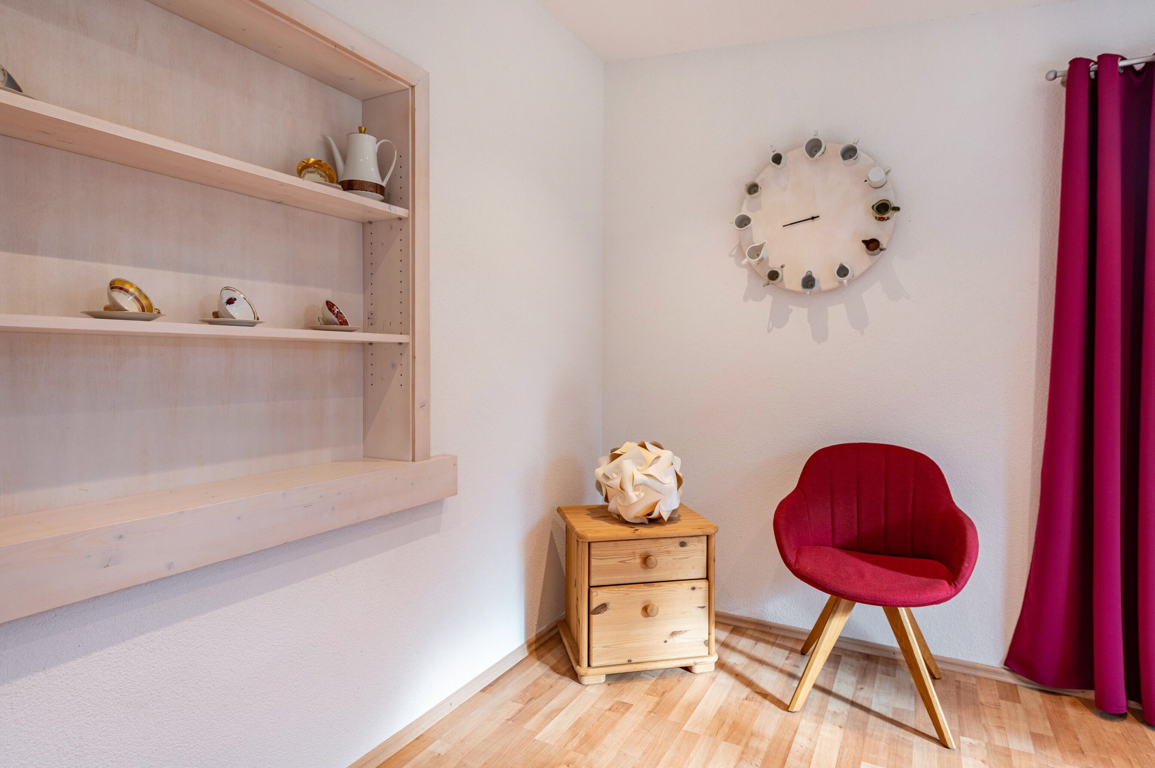 A room with a wooden shelf unit, a small wooden nightstand with a decorative vase, a red chair, and a wall clock.