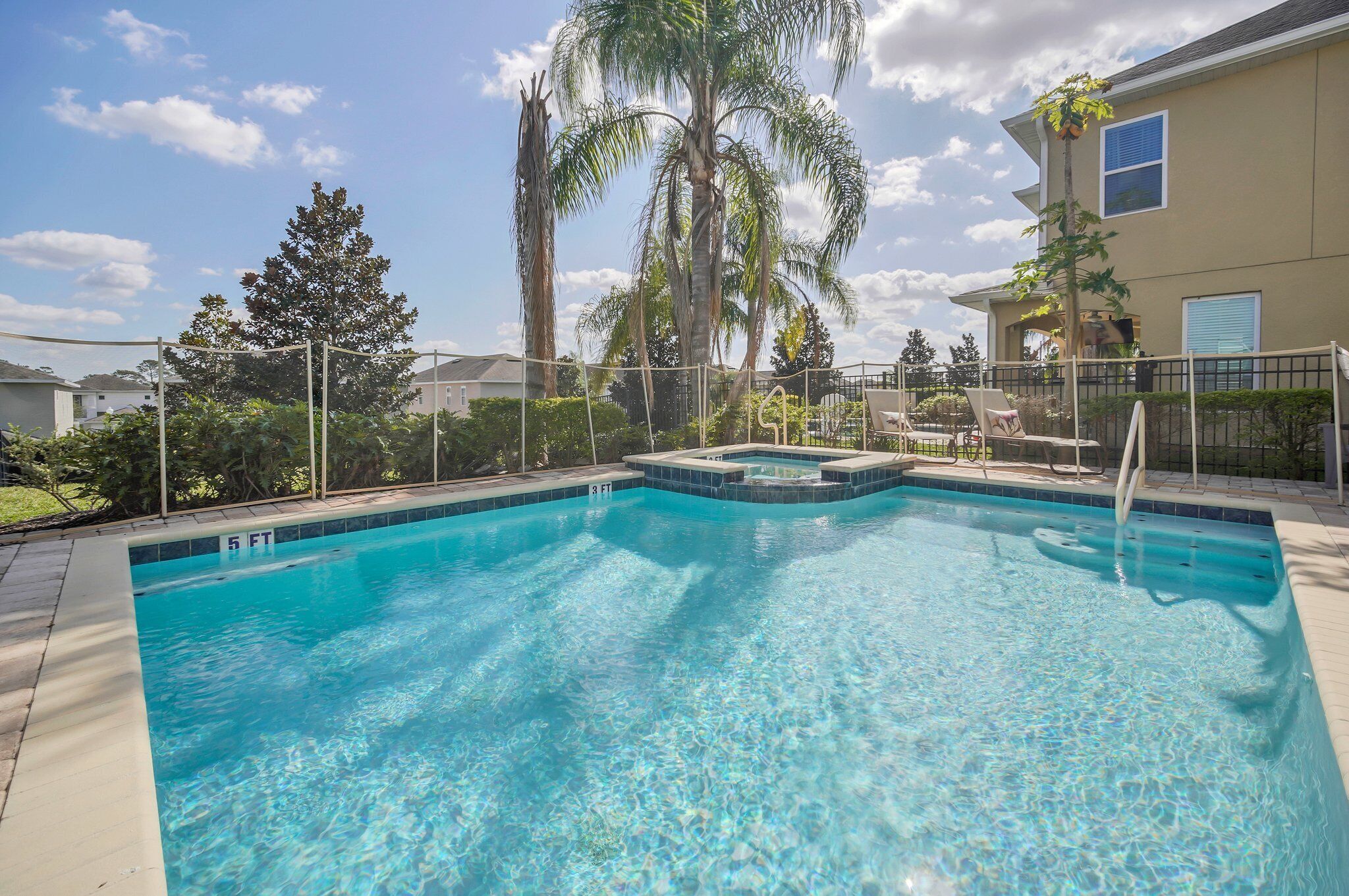 A swimming pool with a 5 FT depth marker, surrounded by a fence and palm trees.