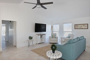 A living room with a ceiling fan, a flat-screen TV, a sofa, two armchairs, a small side table, and a plant.