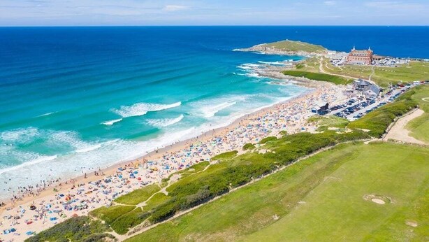 Une vue aérienne d’une plage où l’on aperçoit de nombreux parasols et des personnes, un bâtiment avec un toit rouge et l’océan en arrière-plan.
