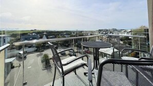 Un balcon doté d’une balustrade en verre, donnant sur un parking et offrant une vue lointaine sur des maisons et la verdure.
