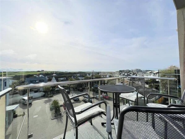 A balcony with a glass railing overlooking a parking area and a distant view of houses and greenery.