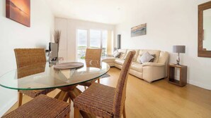 Un salon moderne avec une table à manger en verre, des chaises en osier, un canapé blanc et une petite table d’appoint.