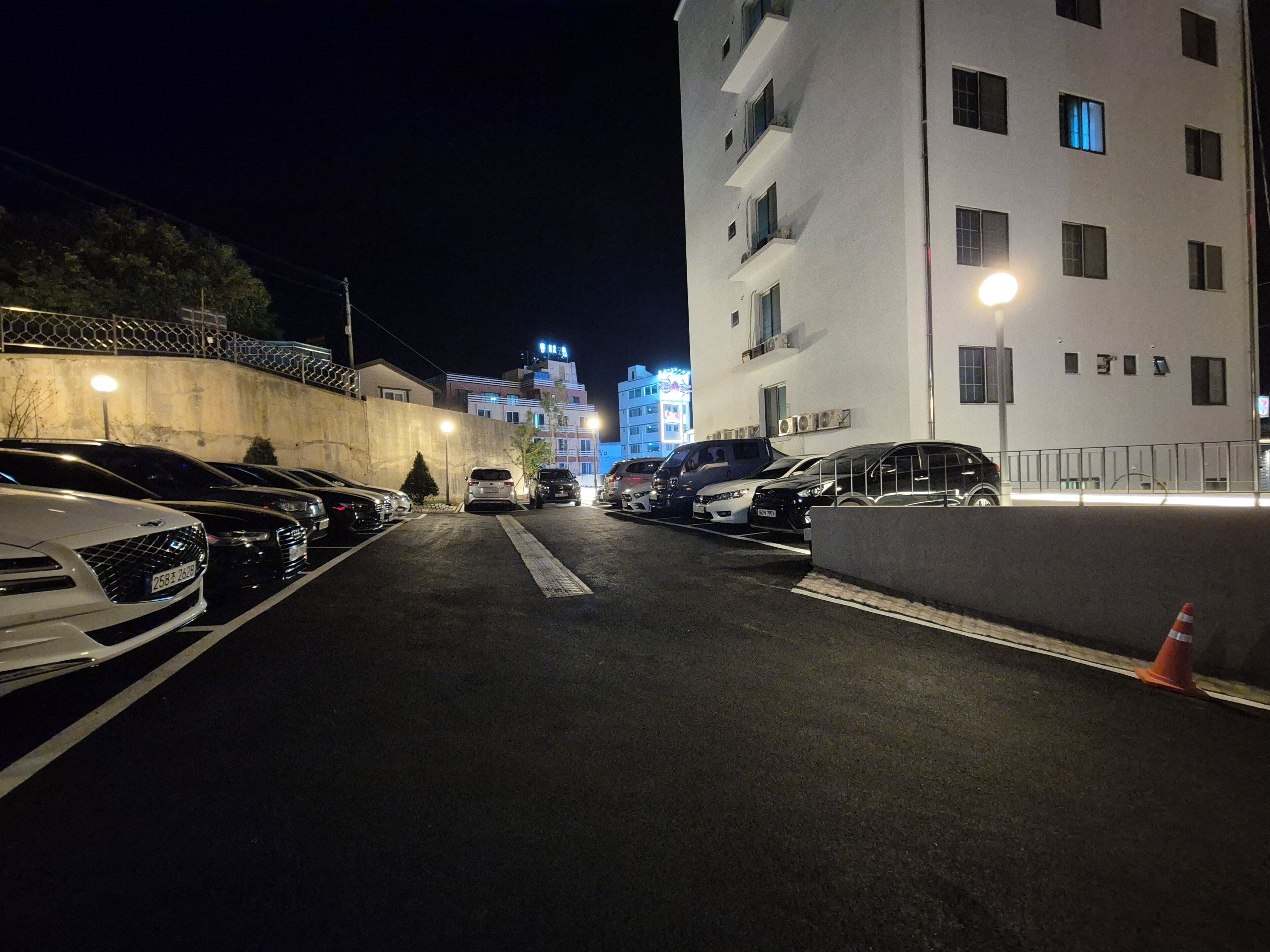 A nighttime parking lot with cars parked along the sides, a building with multiple floors and windows, and a brightly lit sign in the background.