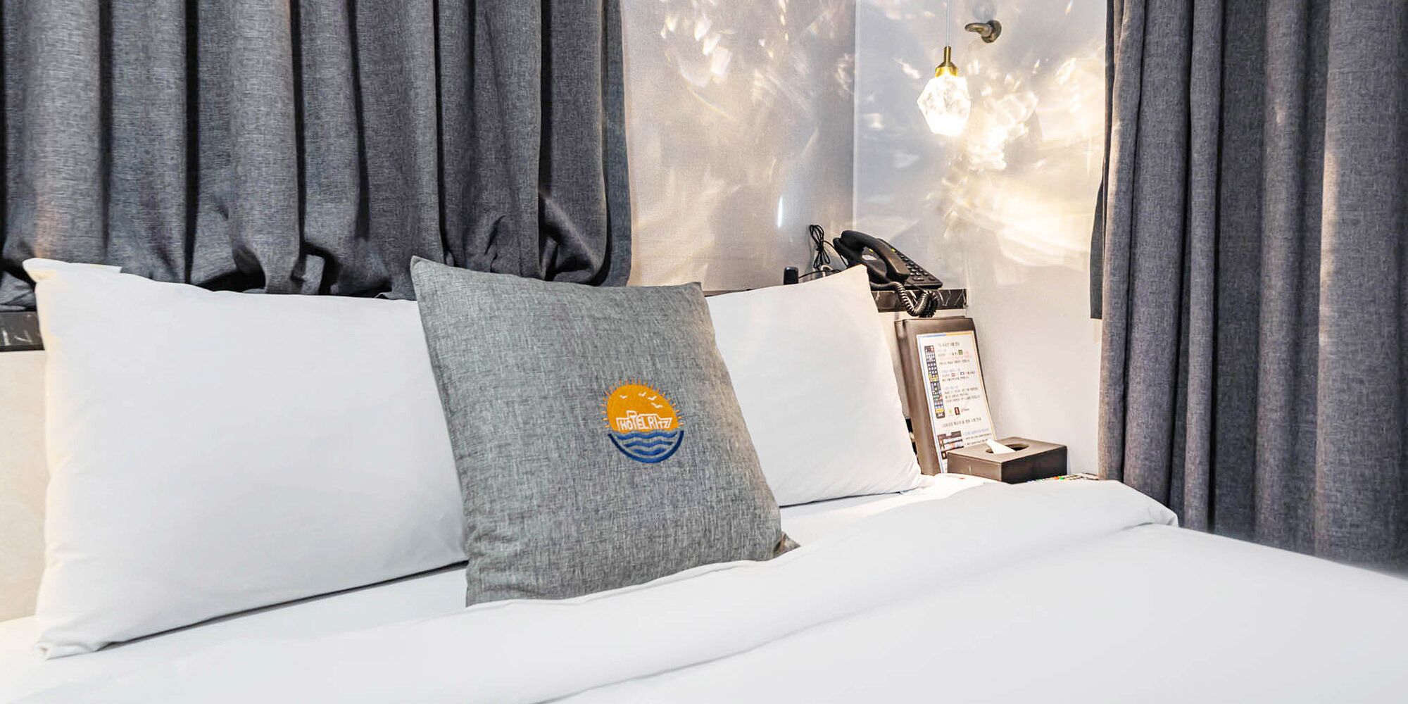 A hotel room with a bed, grey curtains, a nightstand with a telephone, and a wall-mounted light fixture.