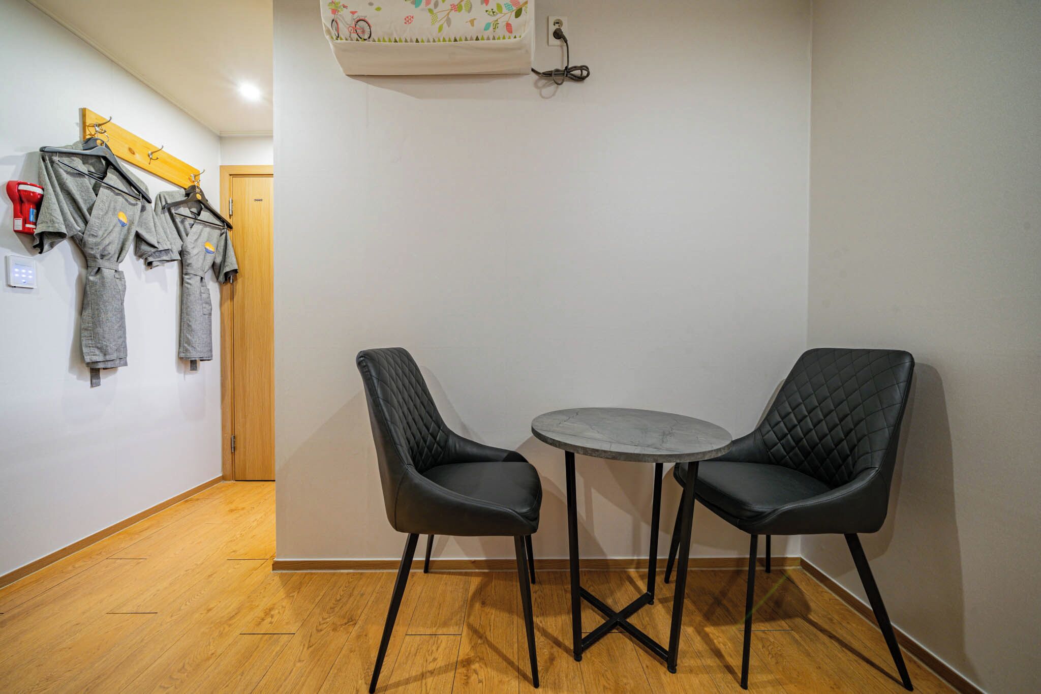 Ein Zimmer mit zwei schwarzen Stühlen um einen kleinen runden Tisch, Holzboden und einem an der Wand befestigten Kleiderhaken mit mehreren Jacken.