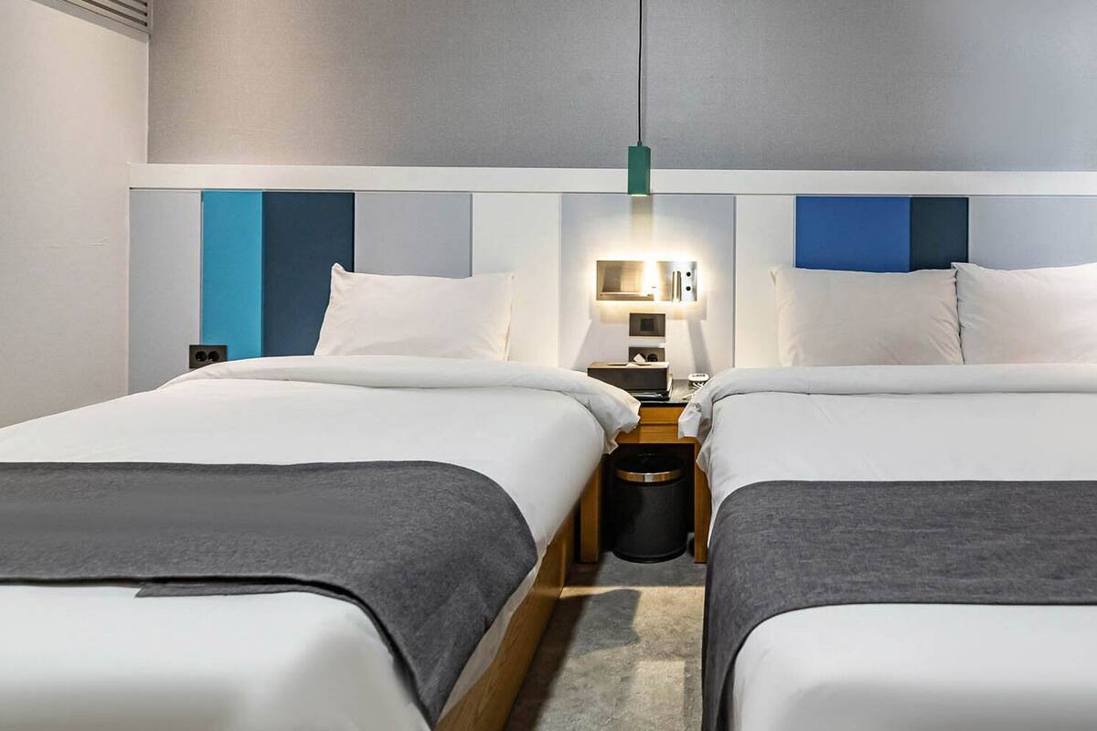 A hotel room with two beds, a nightstand, a lamp, and a trash bin.