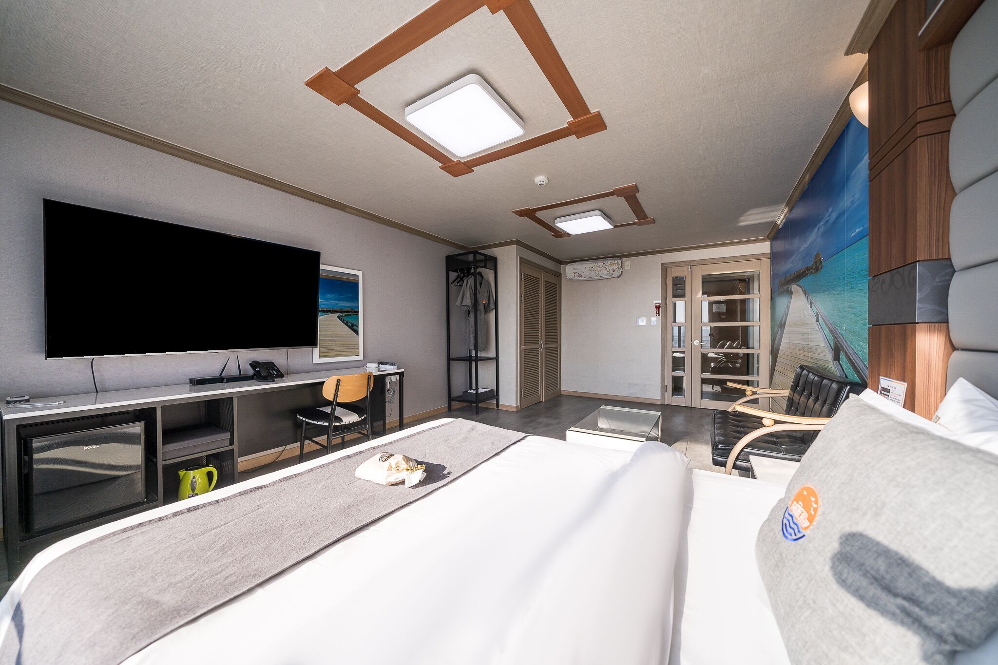 Una habitación de hotel moderna con una cama grande, un escritorio con televisor de pantalla plana, una zona de estar con una mesa y sillas, y vista a un barco por la ventana.