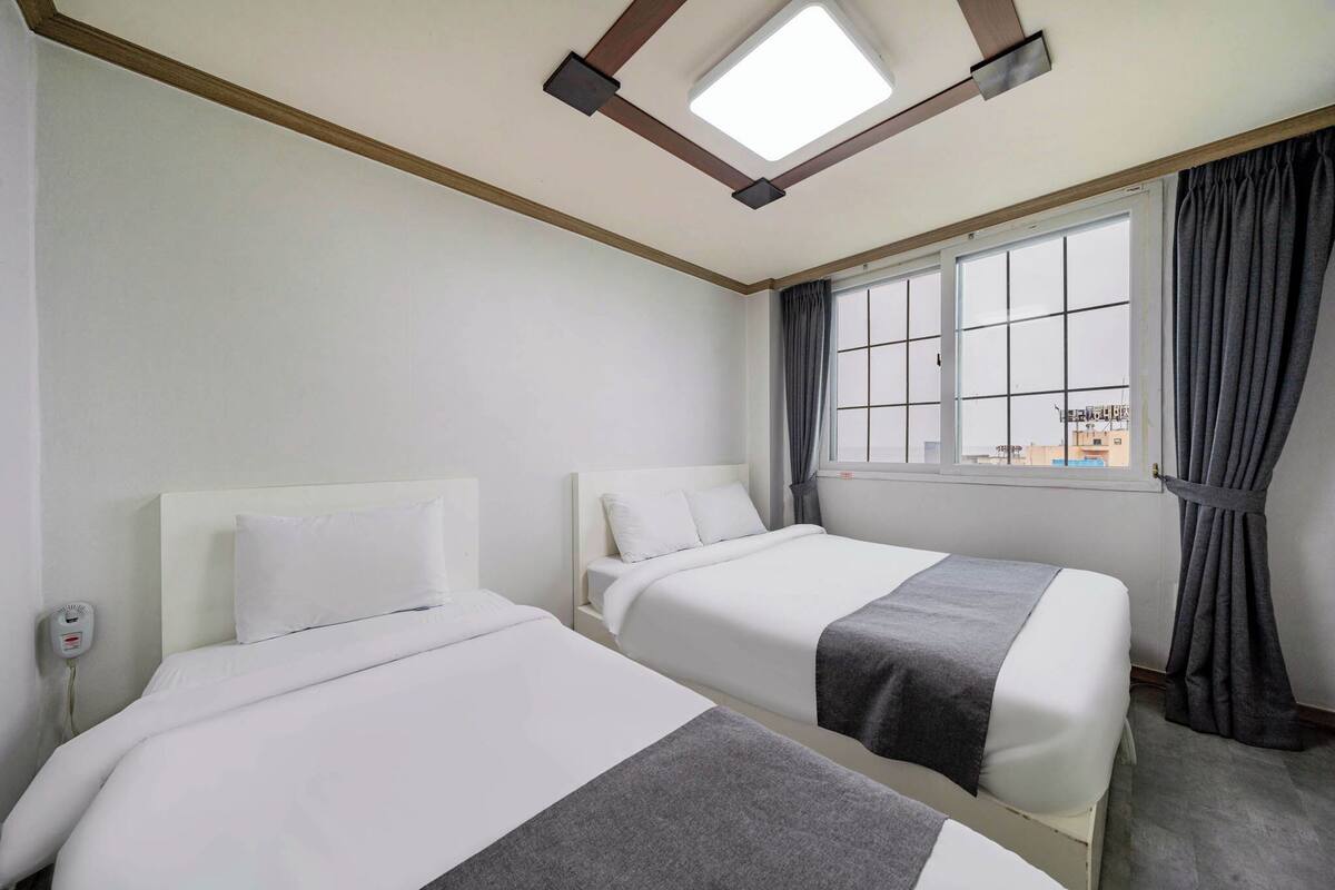 A hotel room with two single beds, a window with curtains, a ceiling light, and a wall-mounted phone.