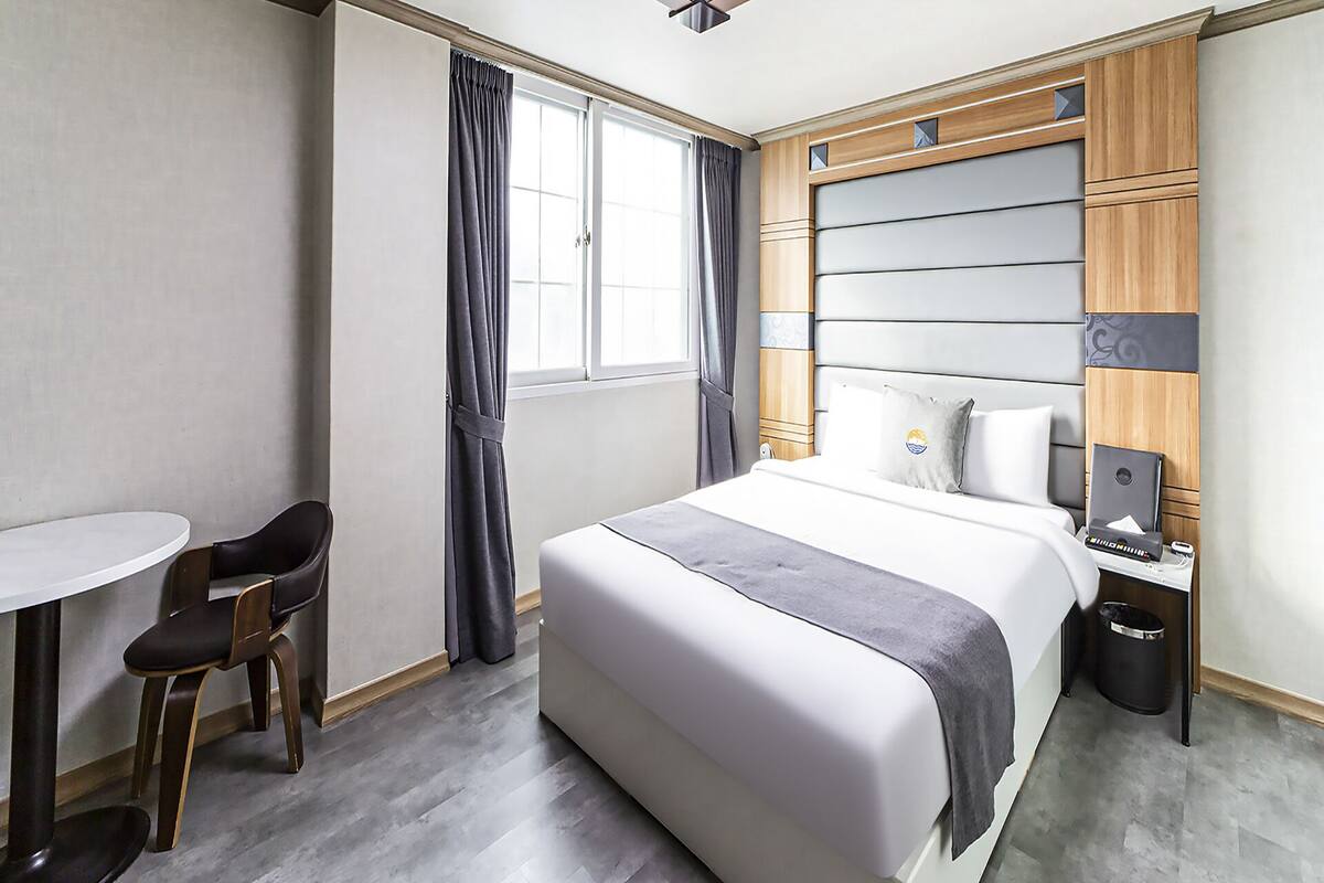 A hotel room with a bed, a desk, two chairs, a window with curtains, and a trash bin.