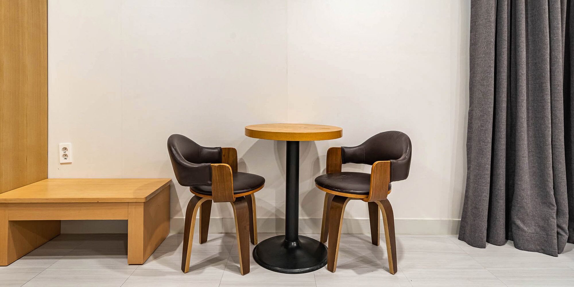 A small, modern room with a round wooden table and two chairs, a wooden bench, and a grey curtain.
