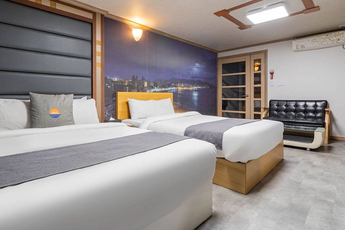 A hotel room with two beds, a cityscape mural, a wooden headboard, and a black leather sofa.