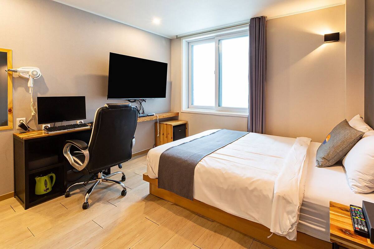 A hotel room with a bed, a desk with a computer, a TV, and a window with curtains.