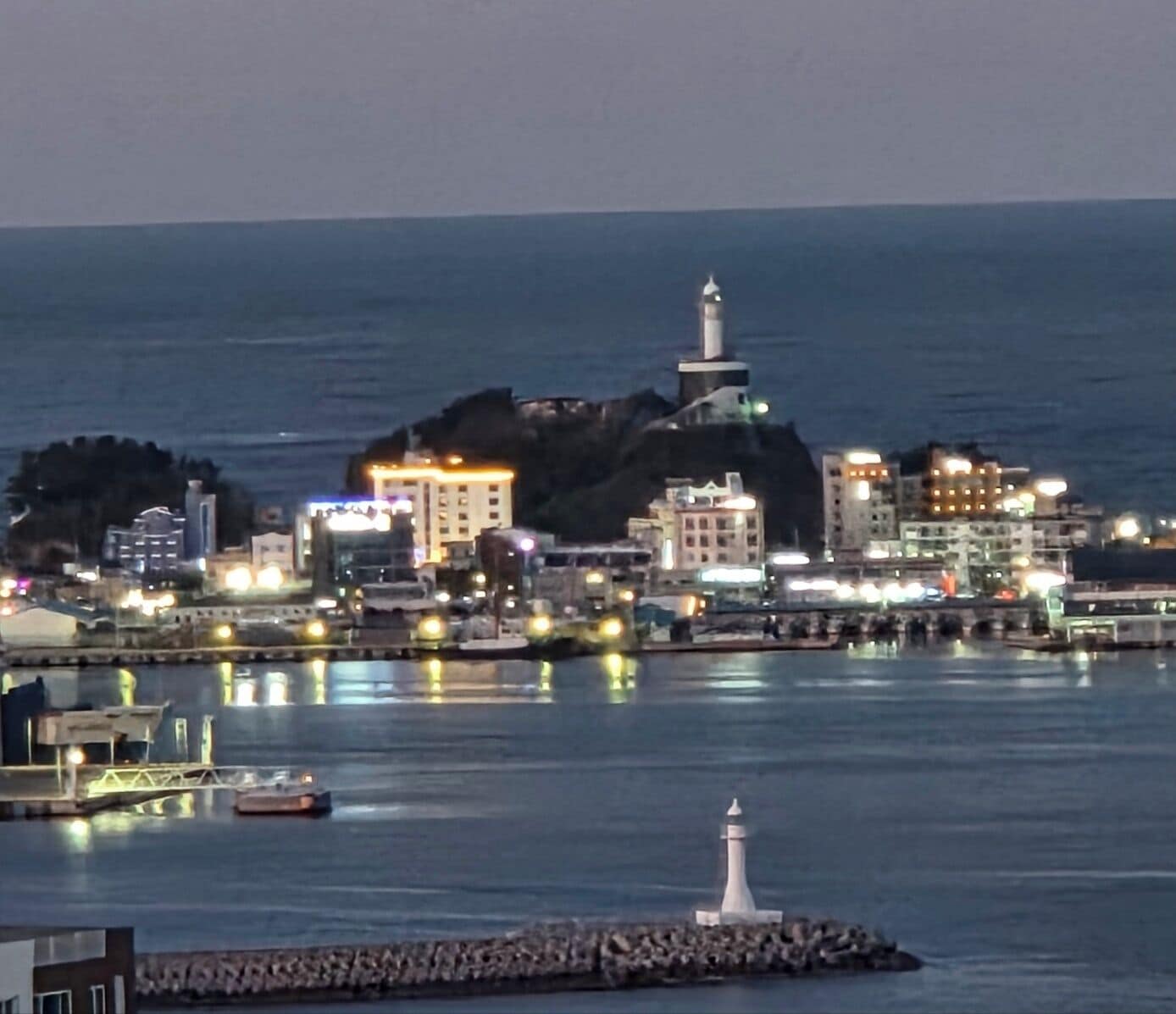 A coastal cityscape with a prominent lighthouse, buildings, and a calm sea.