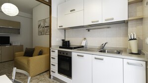 A compact kitchen with white cabinets, a stove, oven, and a sink. There is a yellow armchair and a television in the adjacent living area. - Italianway - Martinetti 21 B (Milan)
