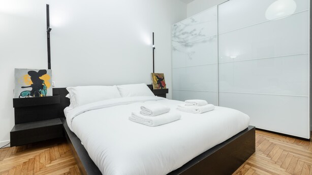A modern bedroom with a bed, white bedding, a black bedside table, and artwork on the wall. - Italianway Comfort - Merlo 1 F (Milan)