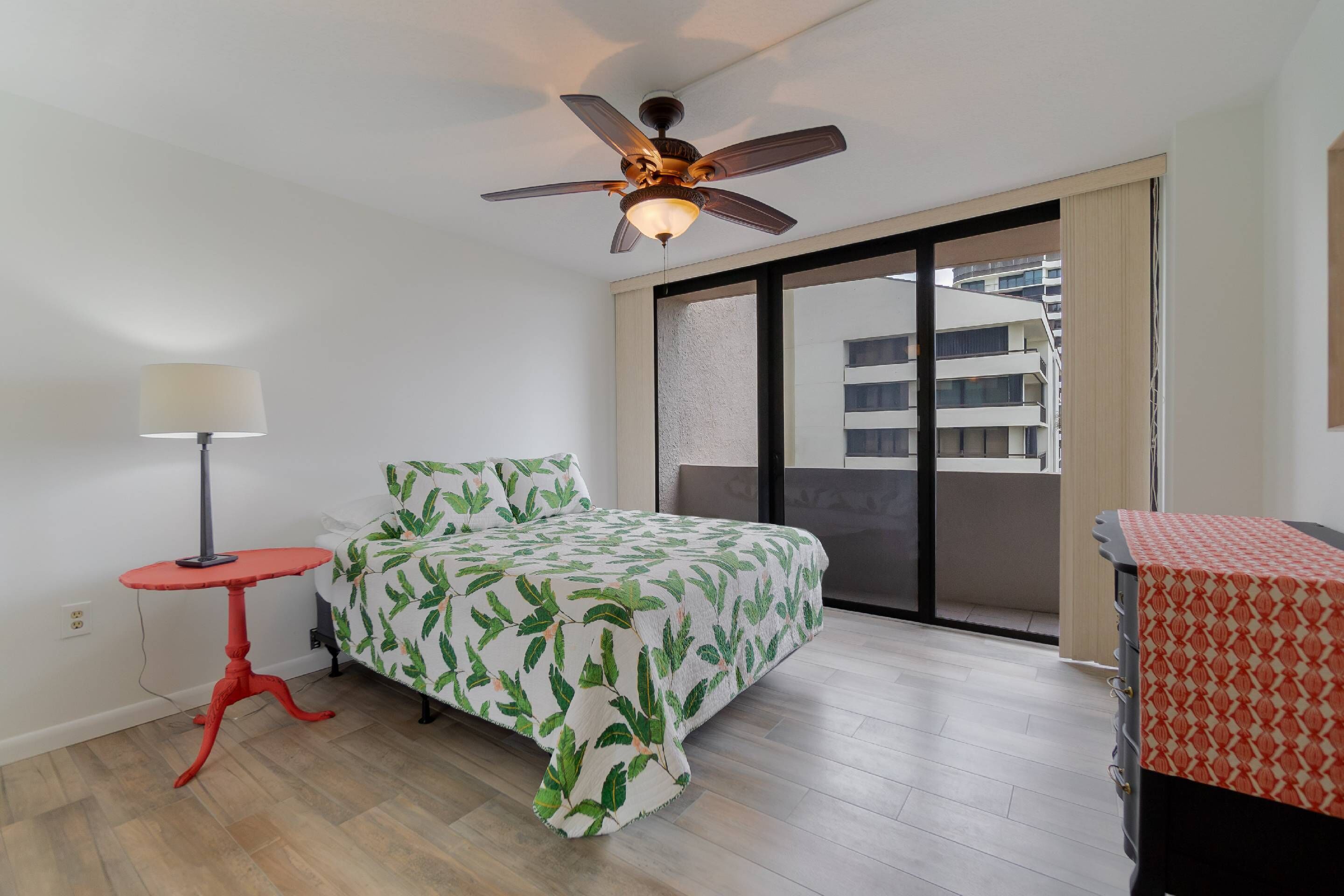 A bedroom with a bed, a ceiling fan, a red table, and a window with a view of buildings.