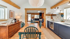 Cocina con vigas de madera, una mesa de comedor, sillas, una lámpara colgante grande y vista al área de estar.