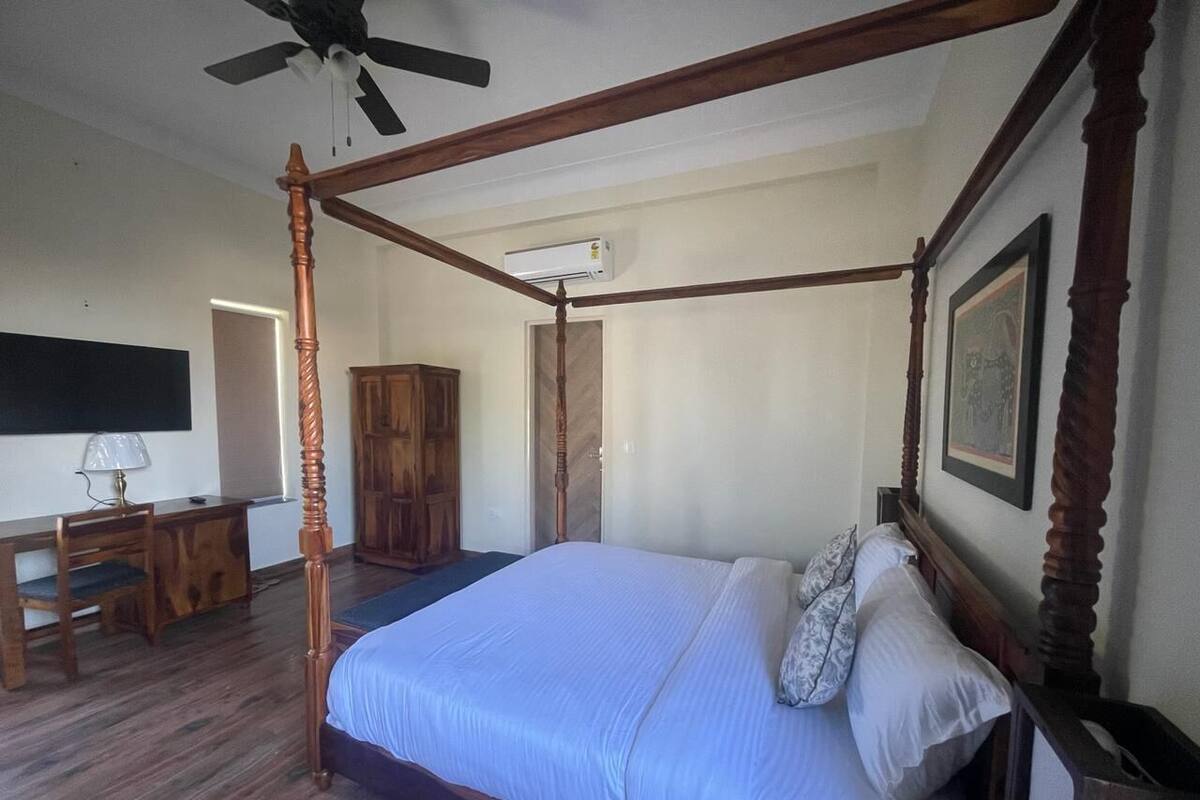 Una cama con dosel de madera y una colcha blanca, un escritorio con lámpara, y un armario en un dormitorio.