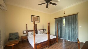 Un lit à baldaquin avec une structure en bois et une literie blanche, un ventilateur de plafond, une table de chevet en bois avec une lampe, et une chaise avec un coussin.