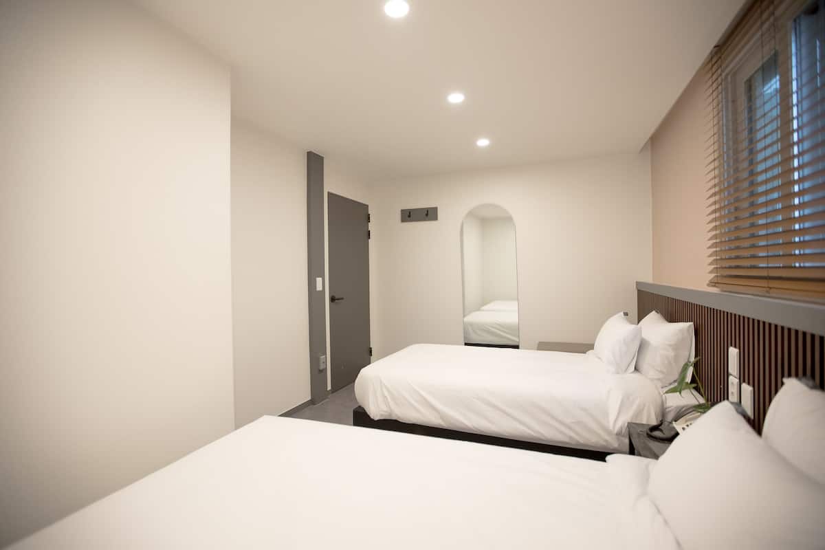 A hotel room with two beds, a mirror, a bedside table, and a window with blinds.
