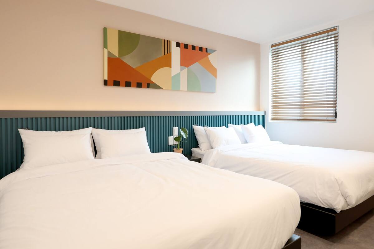 A hotel room with two beds, a teal headboard, a wall-mounted abstract painting, and a window with blinds.