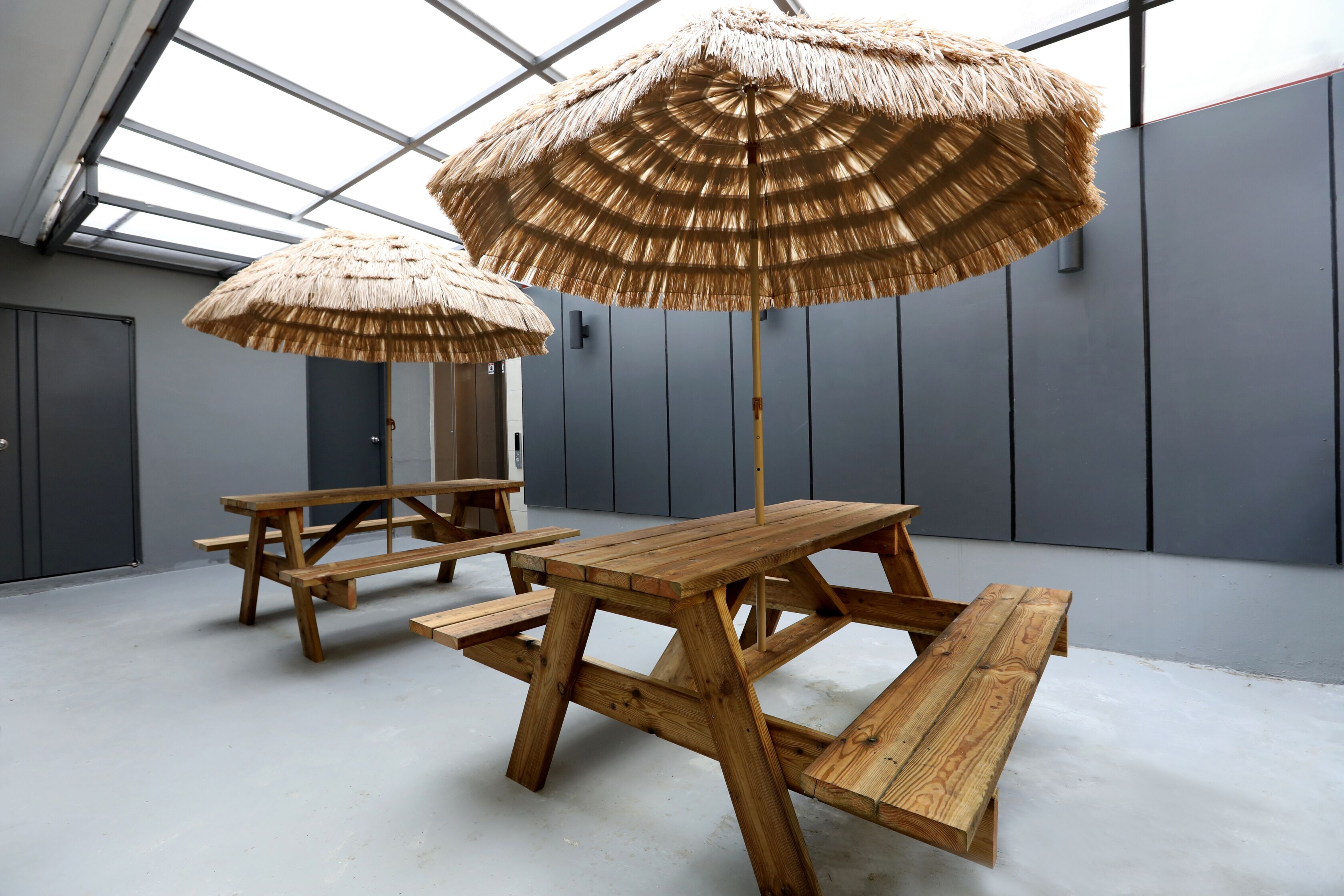 A wooden picnic table with a thatched umbrella in a modern interior space with grey walls and a skylight.