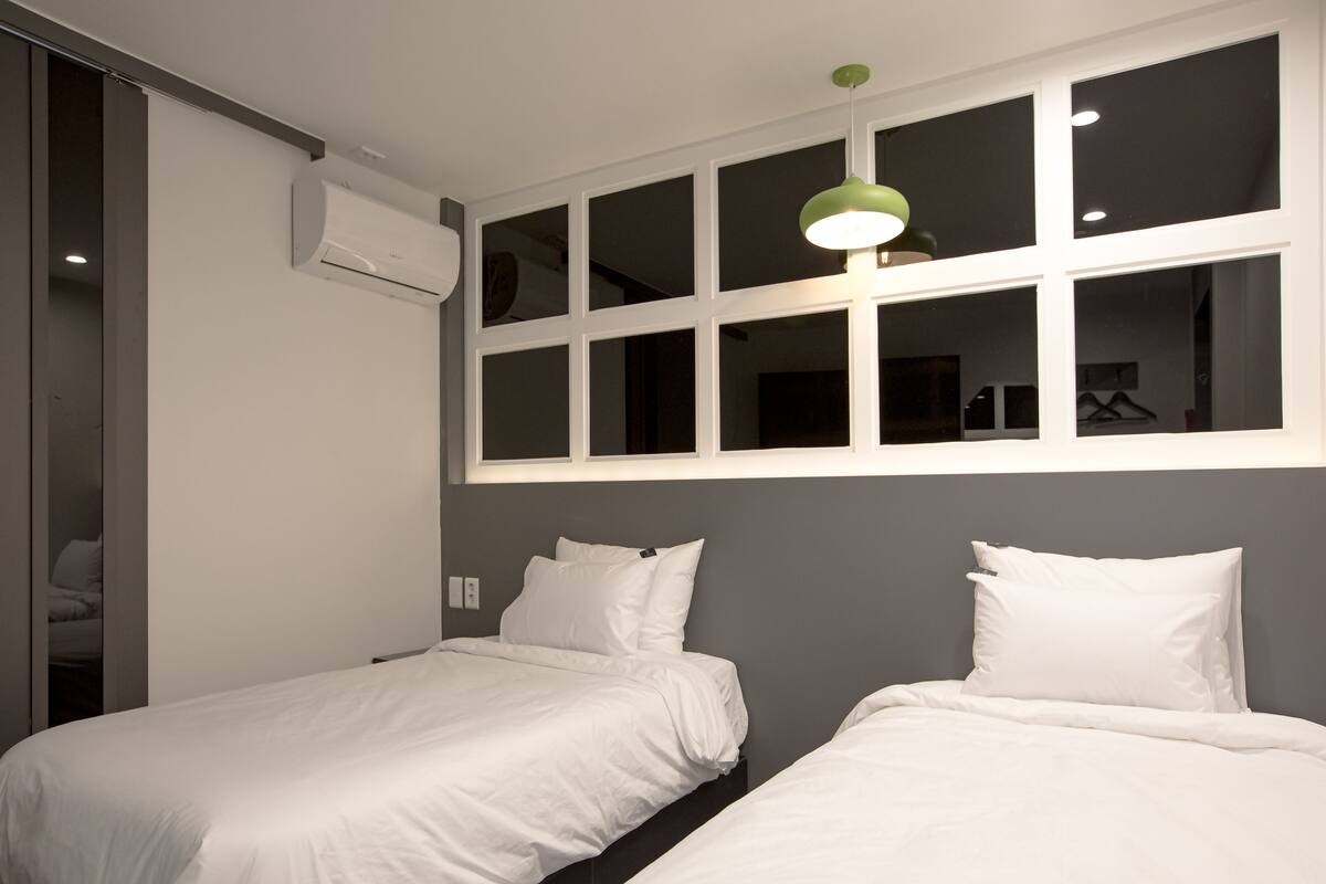 A hotel room with two beds, a large window with multiple panes, and a green pendant light.