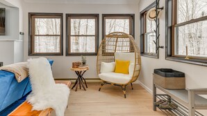 A cozy room with a blue sofa, a white fluffy chair, and a wicker chair with a yellow cushion. There are three large windows with a view of trees, a small wooden table with books and a plant, and a wicker basket on a shelf. - Family-Friendly Big Bass Lake Vacation Rental! (Covington Township)