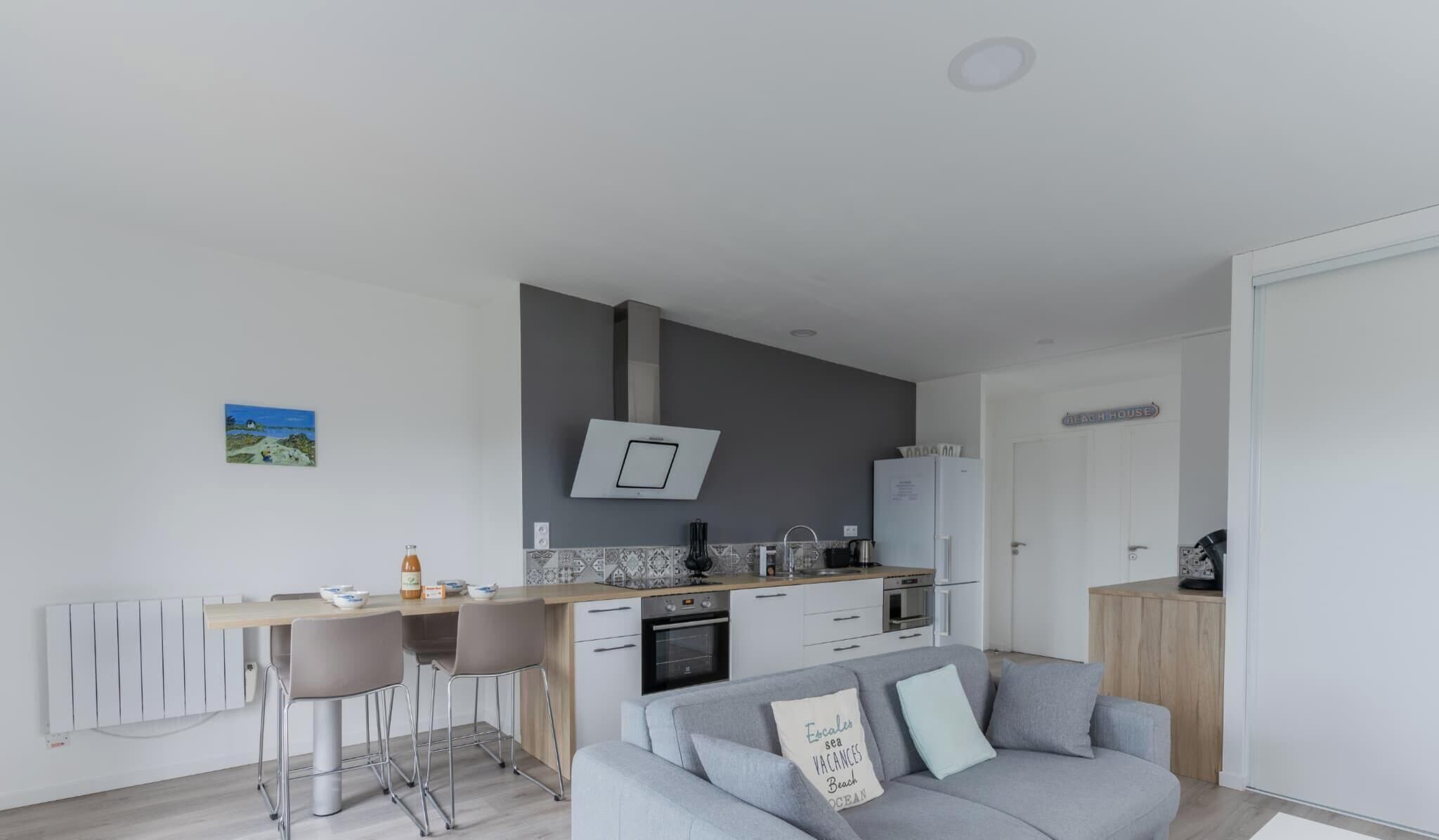 A modern kitchen with a central island, a dining area with bar stools, and a grey sofa with a decorative pillow.