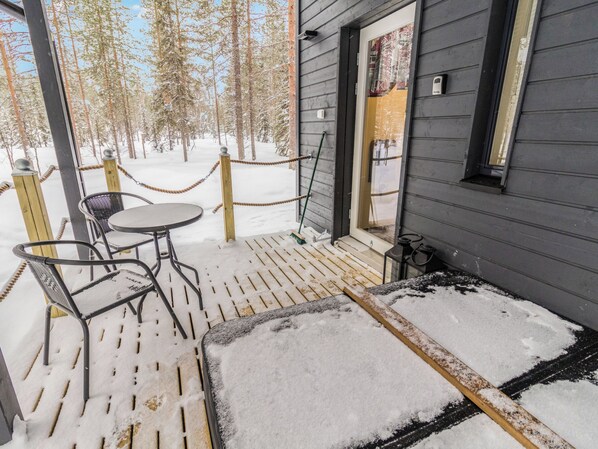 Una terraza de madera con una mesa y sillas de metal, rodeada por una zona nevada y una valla de madera.