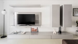 En moderne stue med et fladskærms-tv monteret på en hvid væg, et minimalistisk konsolbord med pynteting, og en gulvlampe.
