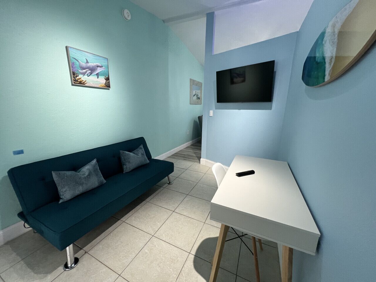 A room with a teal sofa, a white table, and a TV mounted on the wall.